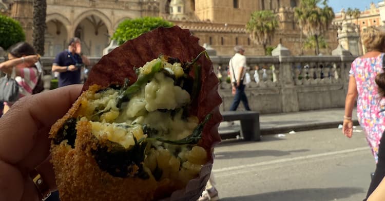 Travelers debating near Palermo Cathedral as a hand reaches for the last arancina